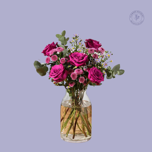 A vibrant floral arrangement called "Amethyst Romance," featuring deep magenta roses, pink daisies, and eucalyptus in a clear glass vase with water.