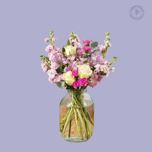 A delicate floral arrangement called "Blush Charm Bouquet," featuring white roses, pink spray roses, alstroemeria, and stock in a clear glass vase with visible stems and water.