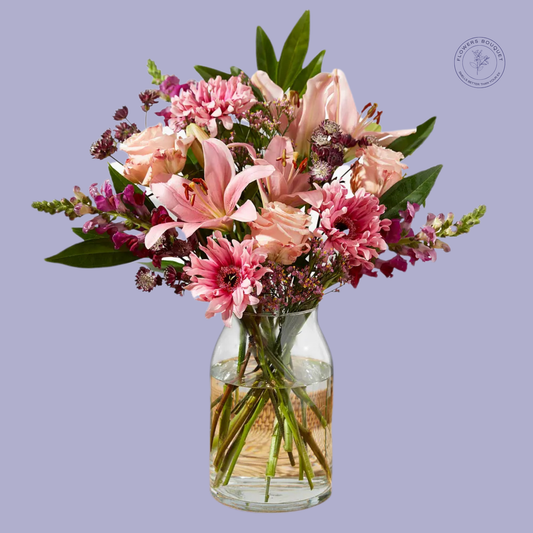 A graceful floral arrangement called "Blush Enchantment," showcasing pink lilies, roses, gerberas, and snapdragons in a clear glass vase with visible water and stems.