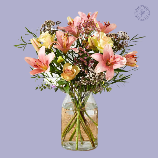 A stunning bouquet called "Blush Lily Elegance," featuring pink lilies, cream roses, gypsophila, and seasonal greenery in a clear glass vase with water.