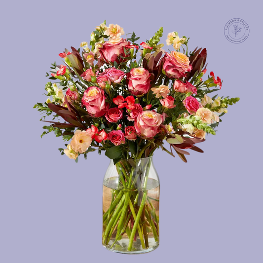 A stunning bouquet called "Blush Symphony," featuring pink roses, peach spray roses, red carnations, snapdragons, and lush greenery arranged in a clear glass vase with visible stems and water.