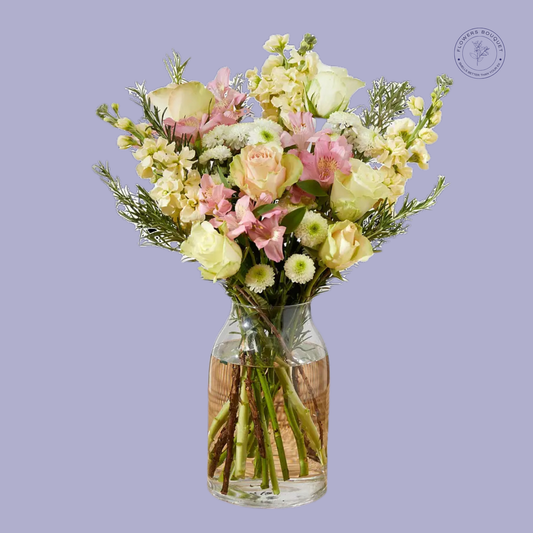 A graceful bouquet called "Golden Grace," featuring yellow roses, pink alstroemeria, stock, and greenery in a clear glass vase with visible water and stems.
