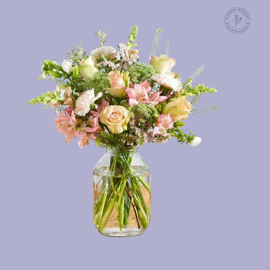 A delicate bouquet called "Pastel Serenity," featuring peach roses, pink alstroemeria, white carnations, snapdragons, and greenery in a clear glass vase with water.