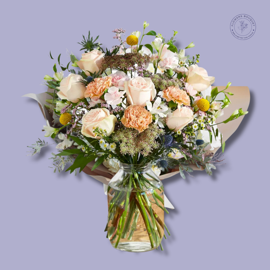 A soft and elegant floral arrangement called "Peach Bloom Harmony," featuring peach roses, carnations, alstroemeria, and wildflowers in a clear glass vase with visible stems and water.