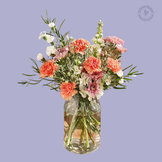 A charming bouquet called "Peach Blossom Delight," featuring peach carnations, pink chrysanthemums, white stock, gypsophila, and greenery in a clear glass vase with water.