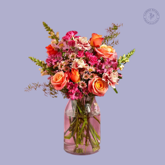 A vibrant floral arrangement called "Sunset Glow Charm," featuring orange roses, pink spray roses, snapdragons, and seasonal blooms in a clear glass vase with water.