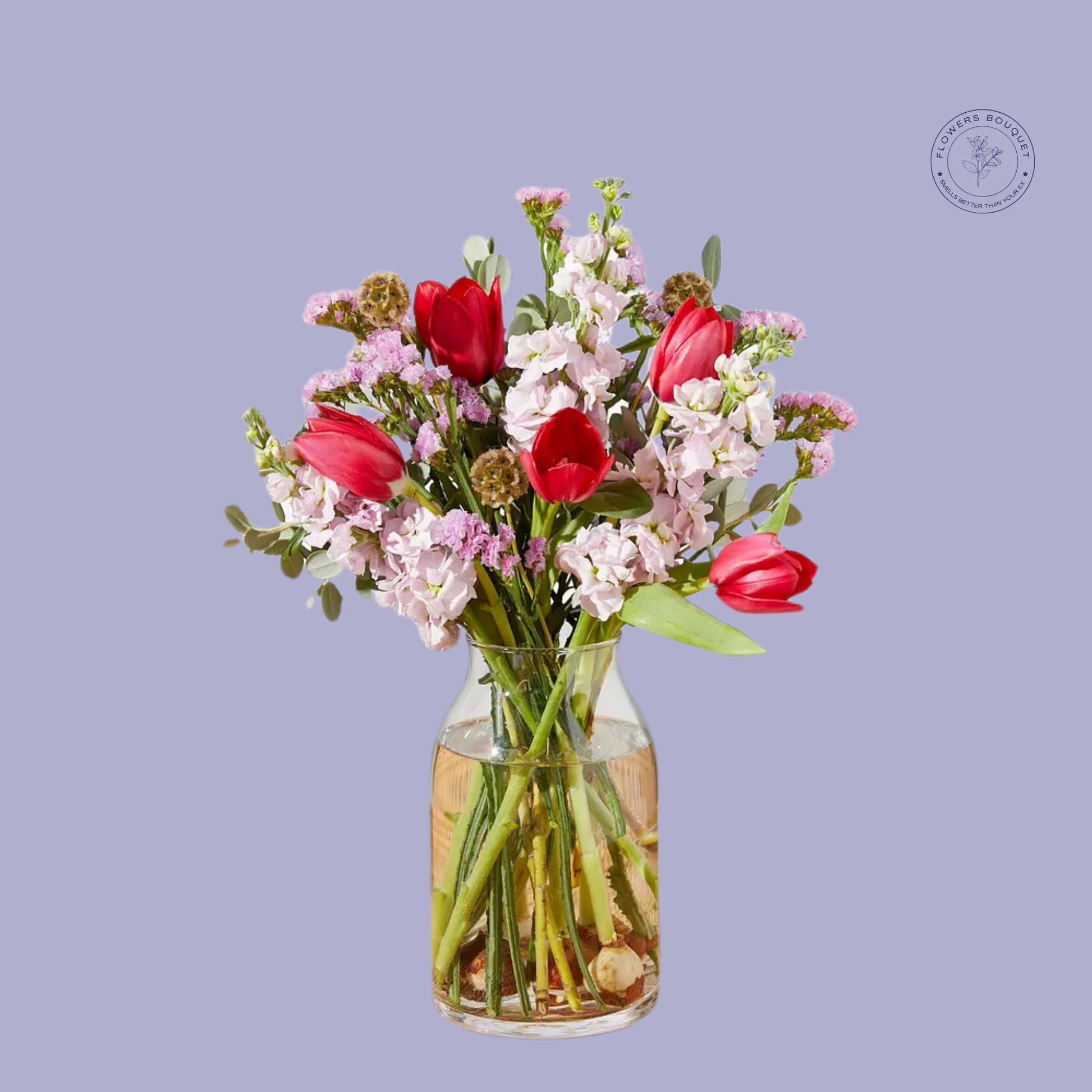 A vibrant floral arrangement called "Tulip Bliss Charm," showcasing red tulips, pink stock, and seasonal accents in a clear glass vase with visible stems and water.