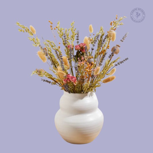 A rustic dried flower arrangement featuring lavender, bunny tails, preserved roses, oats, and poppy pods in a white ceramic vase.